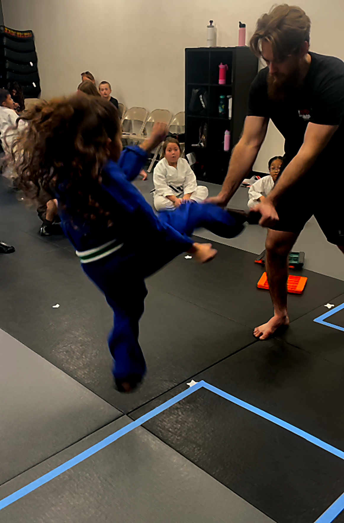 Coach holding a kick pad while a student in a blue gi jumps into a kick