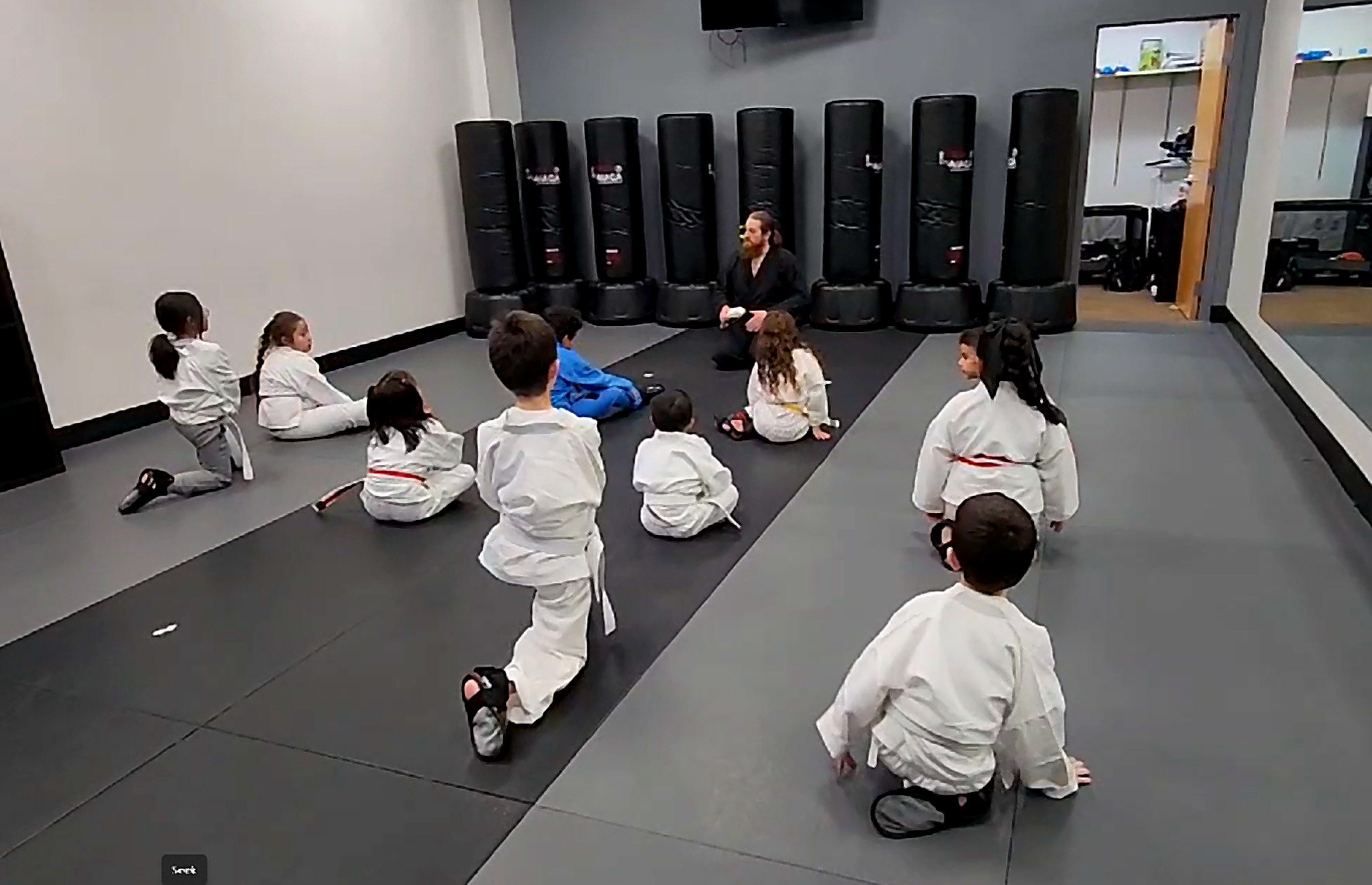 Instructor kneeling on the mats teaching a semi-circle of kids in gis