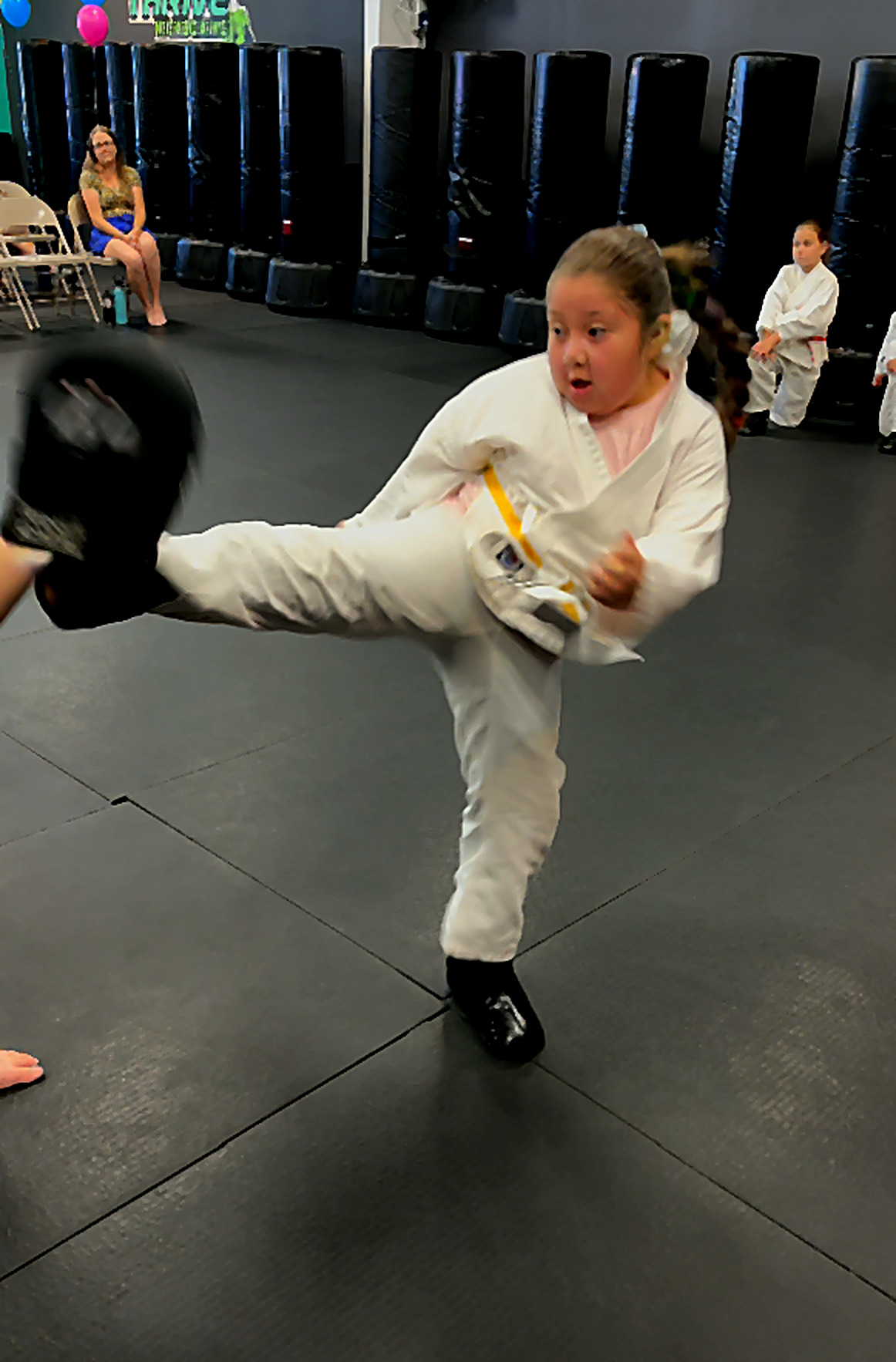 Young girl in a white gi with a yellow belt landing a side kick on a focus pad