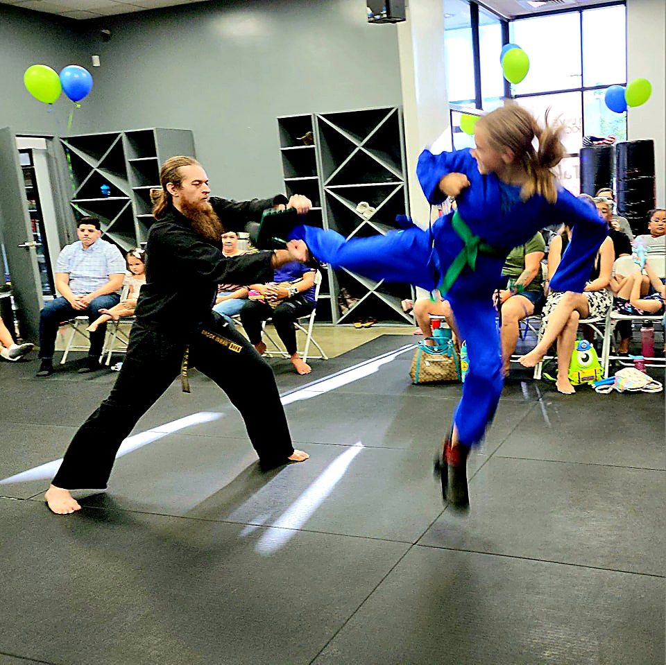 Girl in a blue gi performing a flying kick on a pad held by an instructor during a kids martial arts class in Las Vegas